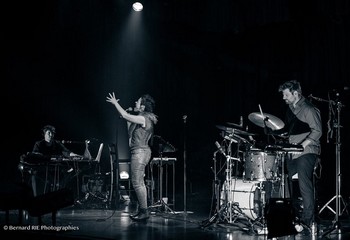 ILIA, LE CONCERT Liège, Cité-Miroir, 26 septembre 2019, Après le disque, le concert. Qui s’ouvre de la même manière : le lancinant Road song, le sautillant Il y a et l’envoûtant Charmeuse de tympans. Trois ambiances pour un bel aperçu de ce que sera la soirée. Sur scène, un batteur et un clavier accompagnent la chanteuse, rejoints par intermittence par un tromboniste. Des musiciens solides pour poser une ambiance propice à l’envolée de la salle dans les sphères de la poésie et de la transe musicale. Le chant d’Ilia est impeccable, bien entendu, malgré un rhume handicapant. Tout sourire, elle nous communique son bonheur d’être là, d’enfin partager ses chansons si longuement mûries, de nous emmener dans son univers à la fois tribal et sophistiqué. L’immense gamme vocale de l’artiste semble défiler (ça mélopée, ça jazze, ça scatte, ça groove), sans ostentation aucune pourtant. Et disons-le, l’émotion est davantage palpable que sur le disque, les petites imperfections scéniques ramenant davantage de vie. Avant de reprendre pour conclure son Il y a accompagnée par le public, Ilia nous offre a cappella une chanson qui l’accompagne depuis des années : La goutte d’eau de Nicole Rieu. Choix surprenant et évident à la fois, tant le timbre de la liégeoise semble façonné pour ce morceau et tant on devine qu’elle s’en sent proche. Ne se termine-t-il pas par : « J'ai quitté la route qu'on m'avait creusée / J'avais trop envie d'être écoutée » ??? (photo Bernard RIE photographies)